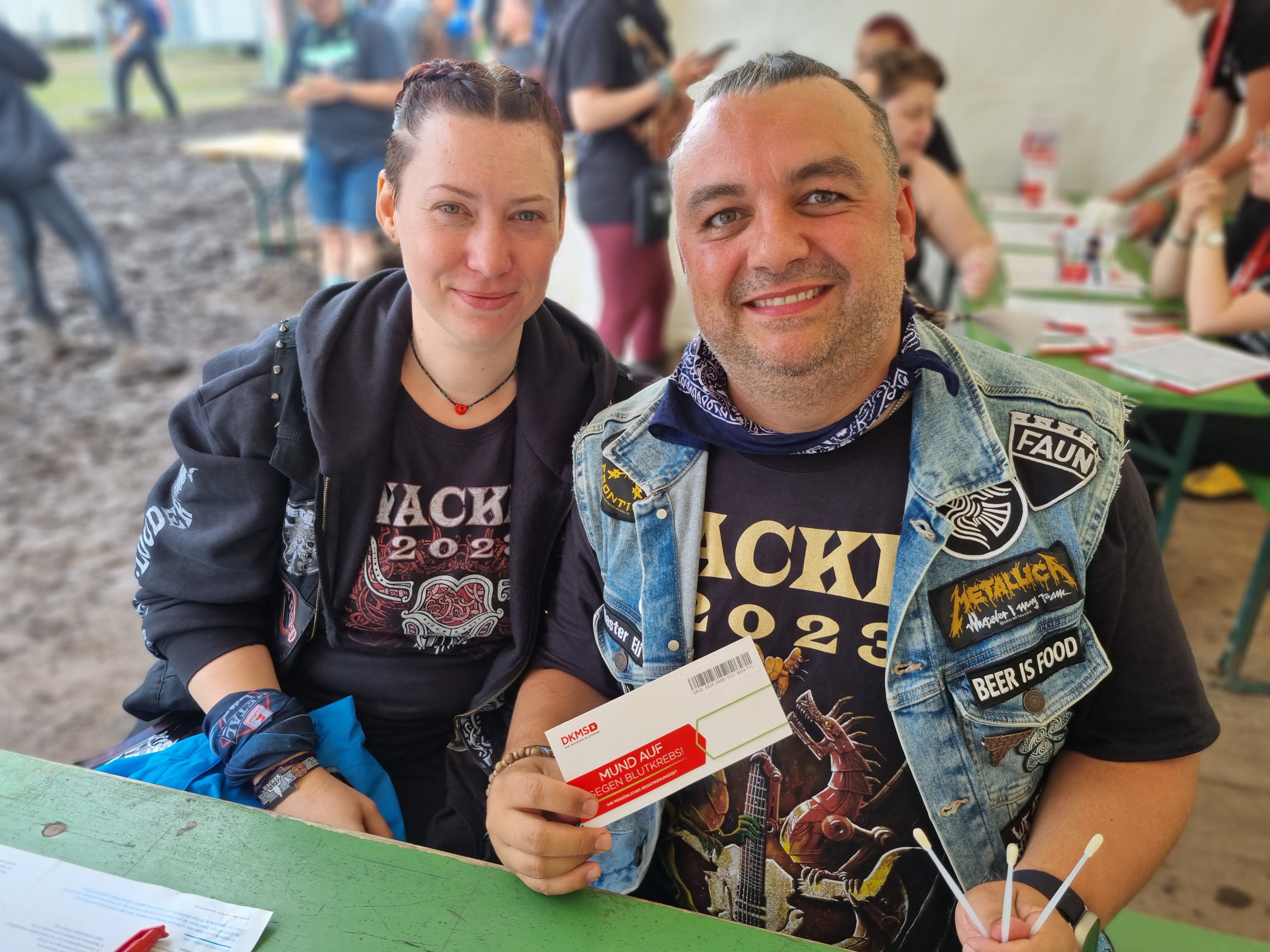 Ein Mann und eine Frau sitzen an einem grünen Tisch in einem Zelt. Beide tragen Wacken 2023-Festivalshirts und Band-Merchandise. Der Mann hält ein DKMS-Registrierungsset in der Hand und lächelt in die Kamera. Vor ihm liegen Wattestäbchen und ein ausgefülltes Formular. Im Hintergrund sind weitere Personen an Tischen und ein matschiger Boden außerhalb des Zelts zu sehen.