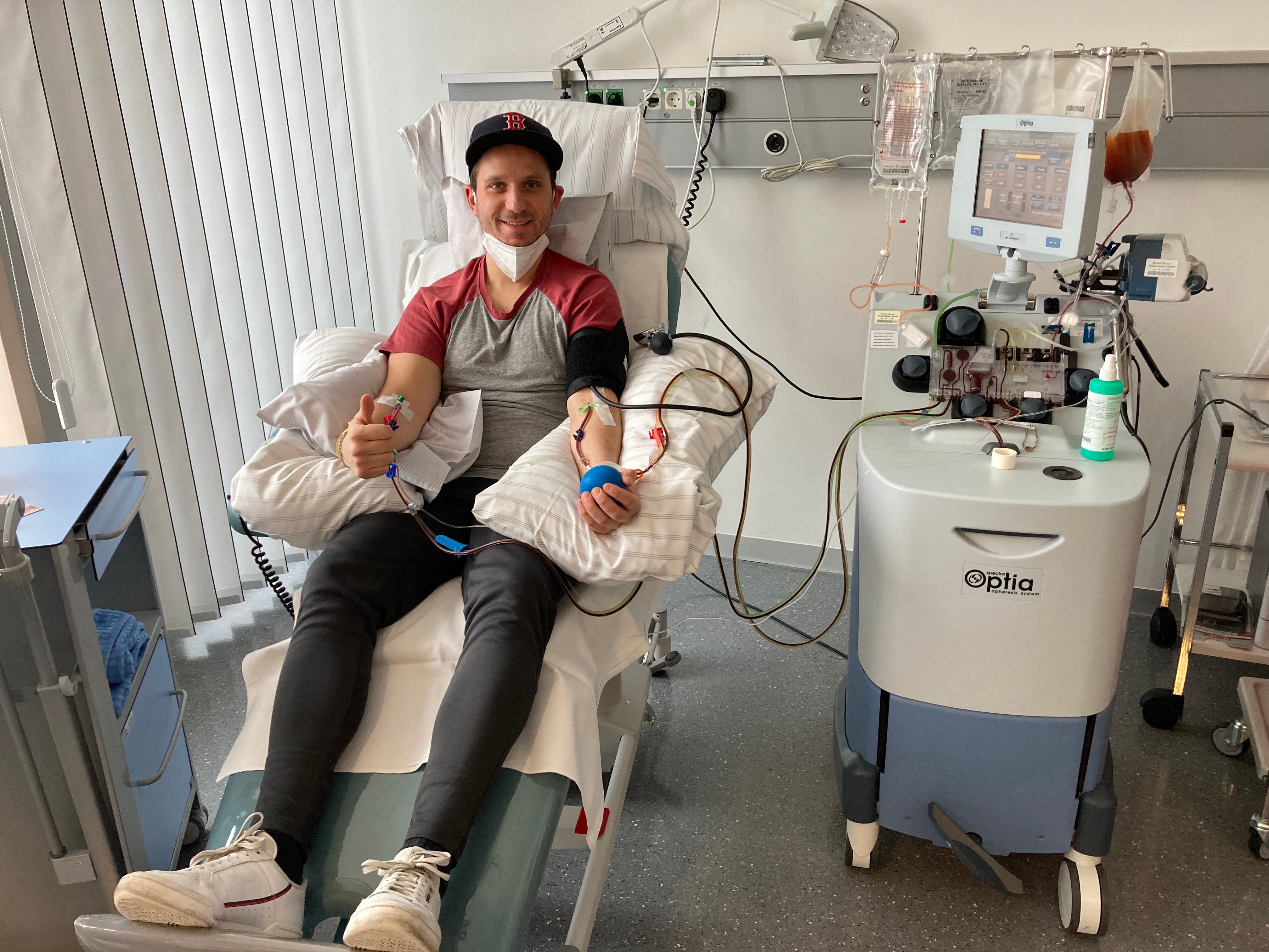 Ein Mann sitzt während einer peripheren Stammzellentnahme in einem Behandlungsstuhl und lächelt in die Kamera. Er trägt eine schwarze Kappe, ein rot-graues Shirt und eine medizinische Maske unter dem Kinn. Beide Arme sind an eine Apheresemaschine angeschlossen. Neben ihm steht die Maschine mit Schläuchen und einem Beutel mit Blutbestandteilen. Der Raum ist hell und medizinisch ausgestattet.

