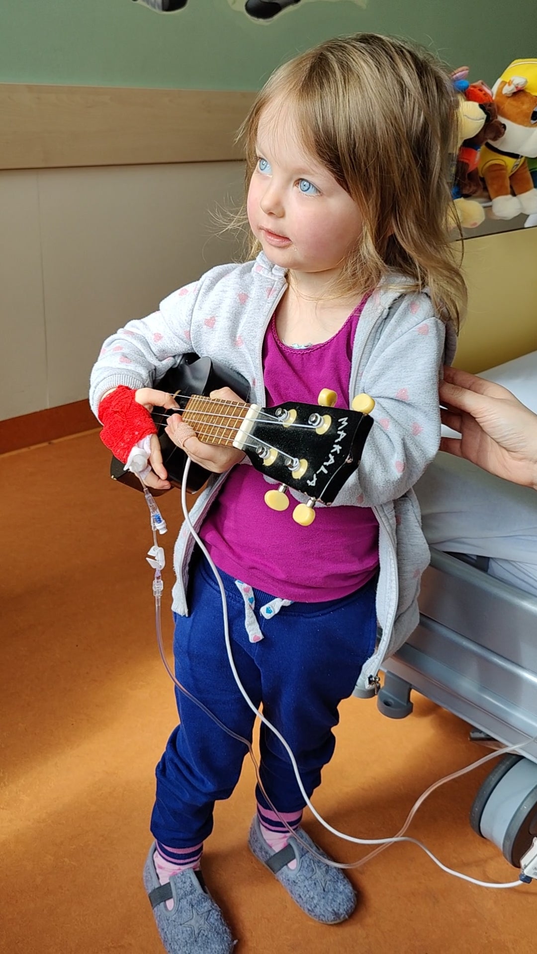 Ein kleines Mädchen steht in einem Krankenzimmer und hält eine kleine Gitarre in den Händen. Sie trägt eine graue Jacke, ein pinkes Oberteil, eine blaue Hose und rutschfeste Hausschuhe. An ihrem rechten Arm befindet sich ein roter Verband mit Zugang, der mit Infusionsleitungen verbunden ist. Neben ihr ist ein Krankenhausbett mit Stofftieren zu sehen.
