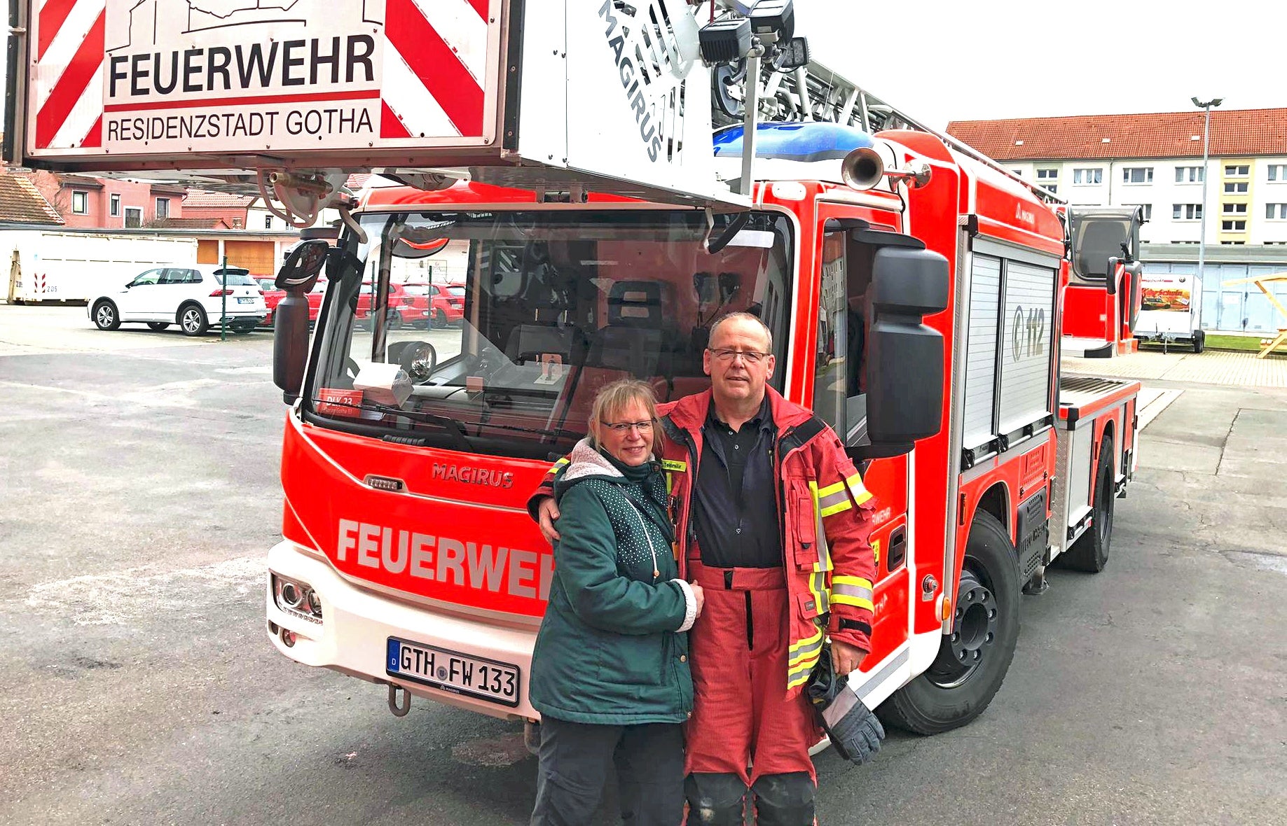 Ein Feuerwehrmann in Einsatzkleidung steht mit einer Frau Arm in Arm vor einem roten Löschfahrzeug der Feuerwehr Residenzstadt Gotha. Beide lächeln in die Kamera. Das Fahrzeug steht auf einem Hof, im Hintergrund sind Wohngebäude zu sehen.