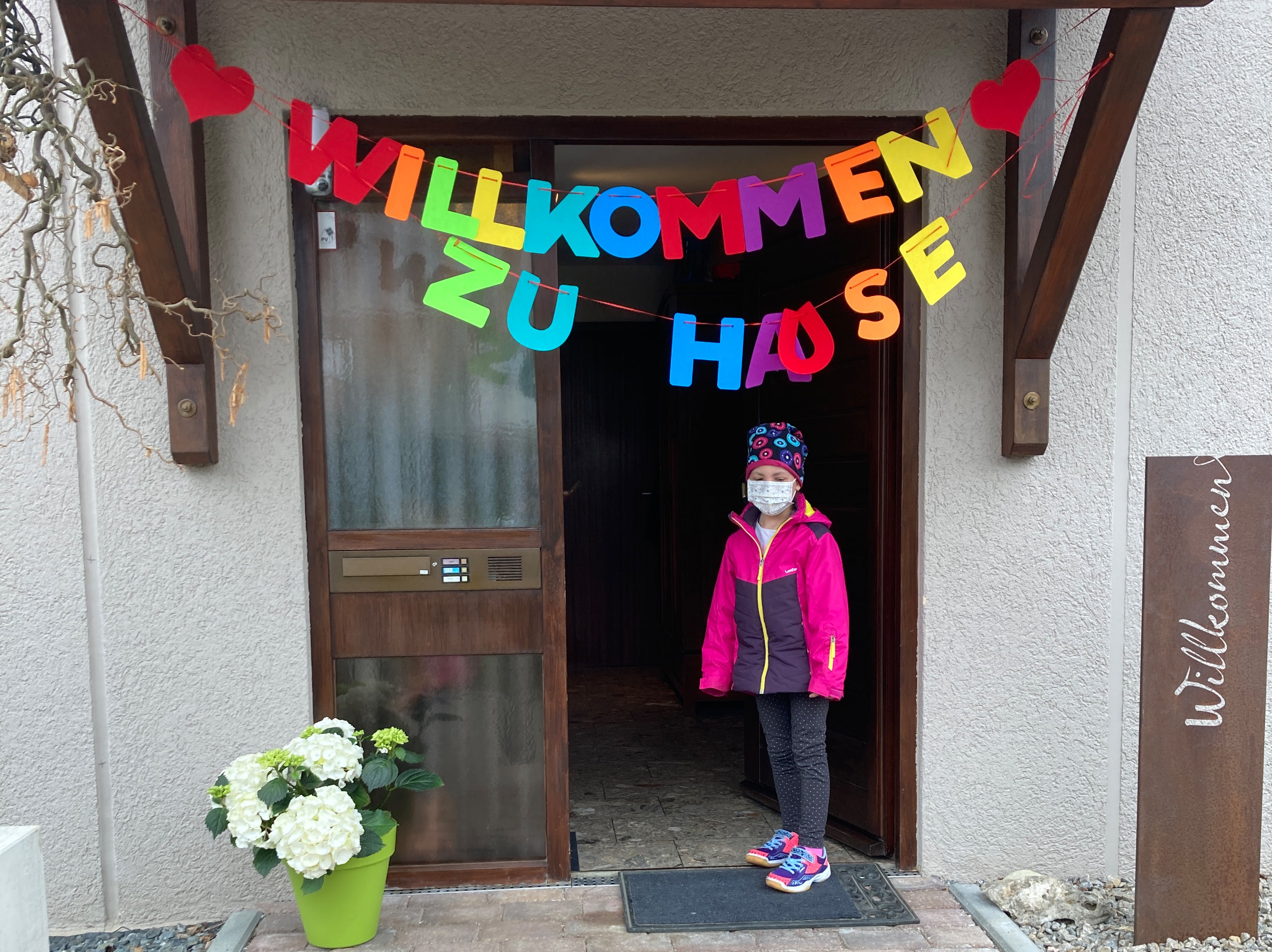 Ein Mädchen steht vor einer Tür, über ihr hängt eine Girlande: Willkommen zu Hause 