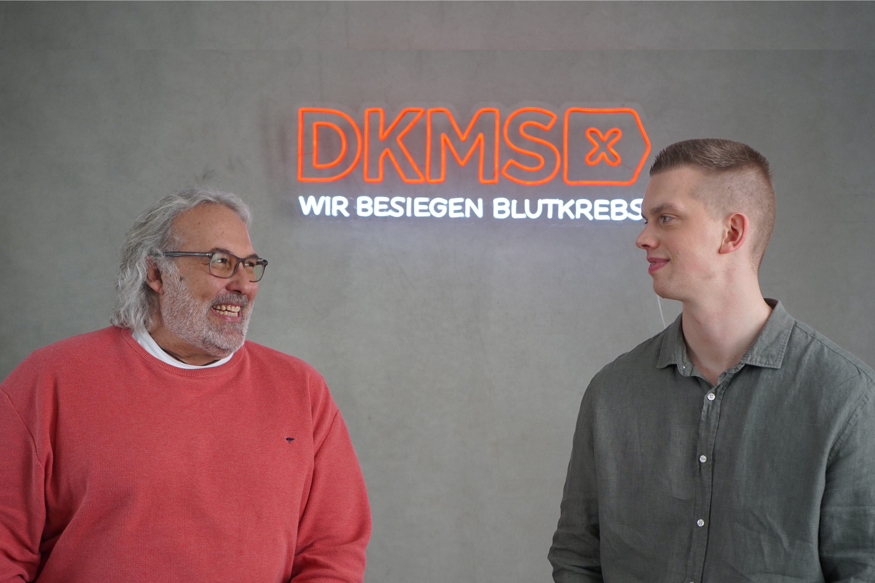 Zwei Männer stehen vor einem Leucht-Schild der DKMS