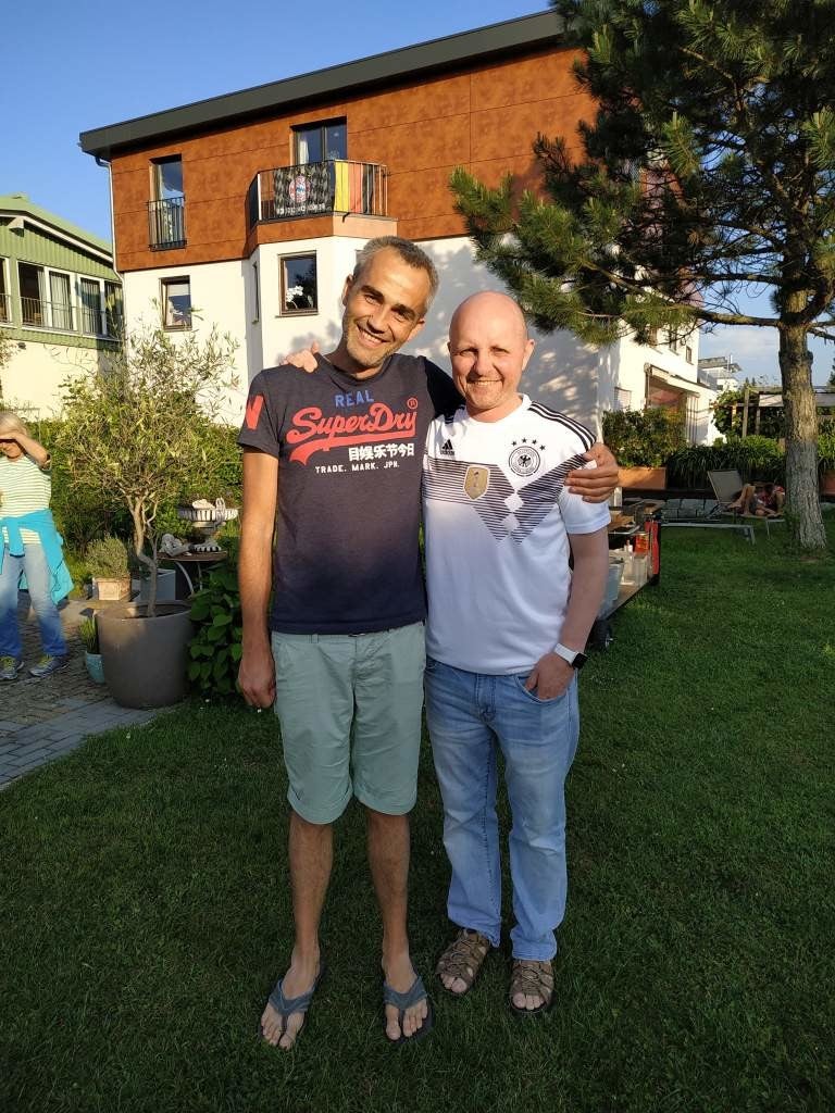 Christian und David stehen in einem Hof. Sie stehen nebeneinander und umarmen sich.