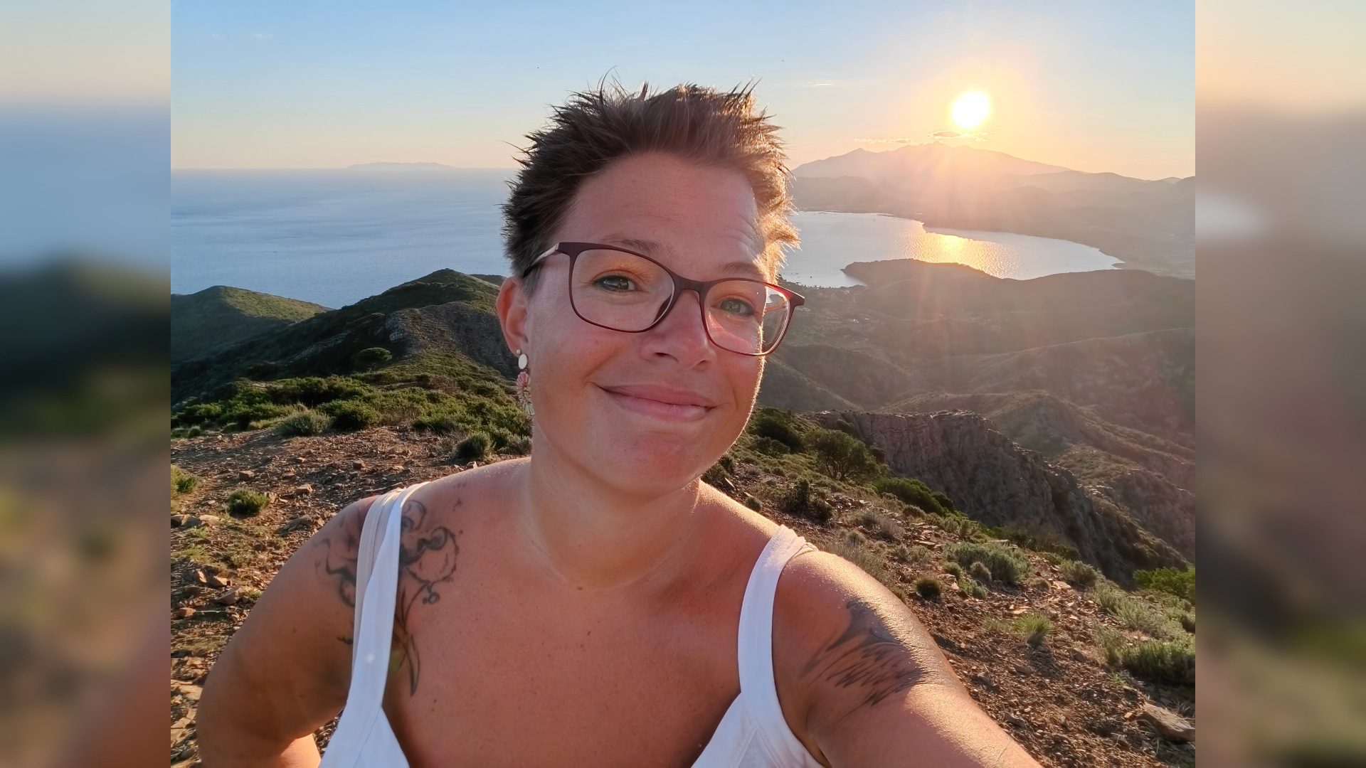 Eine Frau steht in sommerlicher Bekleidung auf einem Berg. Hinter ihr geht die Sonne unter. 