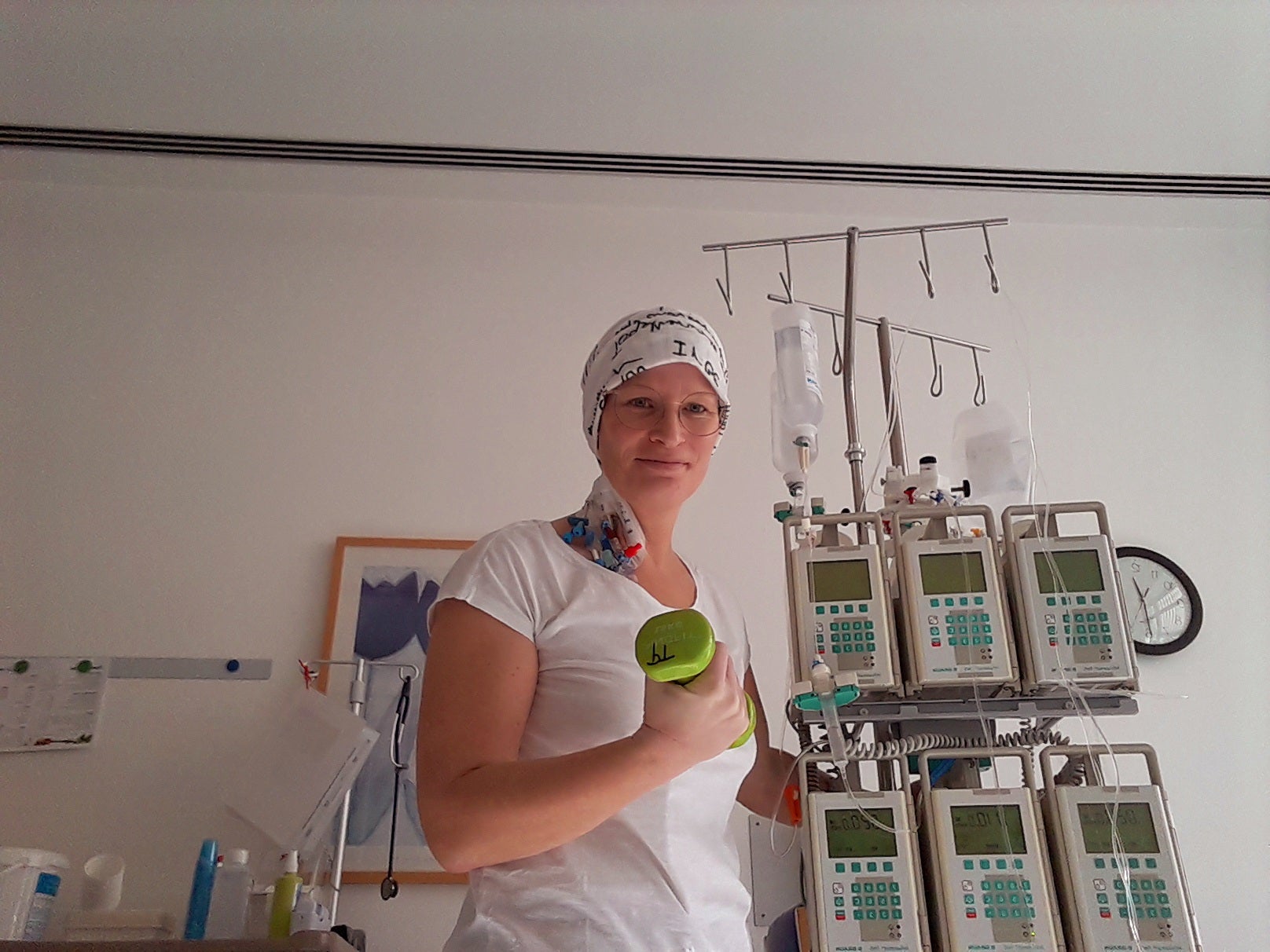 Eine Frau mit Kopftuch steht in einem Klinikzimmer neben einem Infusionsständer mit mehreren Infusionspumpen und Infusionsbeuteln. Sie trägt ein weißes T-Shirt und hält einen grünen Becher in der Hand. Im Hintergrund hängen ein Bild und eine Wanduhr. Die Atmosphäre ist ruhig und klinisch.
