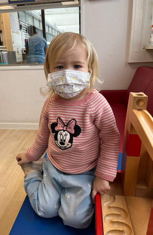 Ein kleines Mädchen mit blonden Haaren in einem rosa-weiß gestreiften Pullover sitzt auf dem Boden. Im Gesicht trägt sie eine Maske und neben ihr ist eine Murmelbahn zu sehen. 