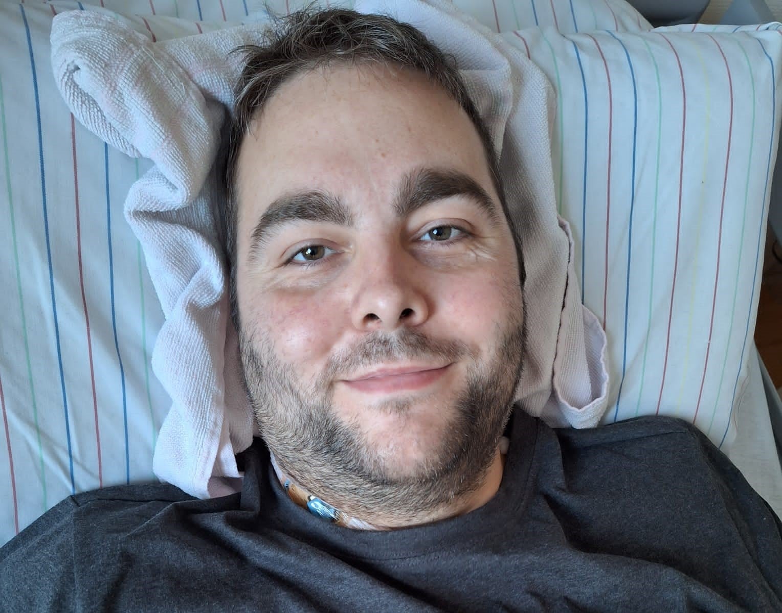 Ein Mann liegt auf einem gestreiften Kopfkissen und blickt in die Kamera. Er trägt ein dunkles T-Shirt, im Halsbereich ist ein medizinischer Zugang sichtbar. Unter seinem Kopf liegt zusätzlich ein weißes Handtuch als Stütze.