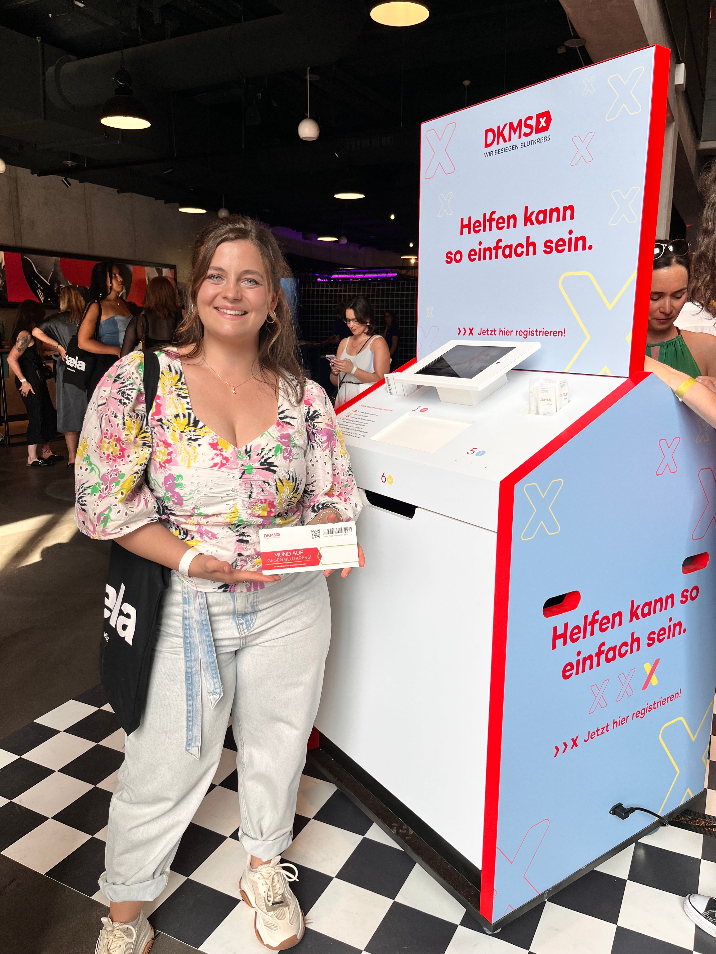 Eine Frau steht lächelnd neben einer DKMS-Registrierungsstation mit der Aufschrift „Helfen kann so einfach sein.“ Sie hält ein Registrierungsset in der Hand. Die Station steht auf einem schwarz-weiß karierten Boden in einem Veranstaltungsraum.