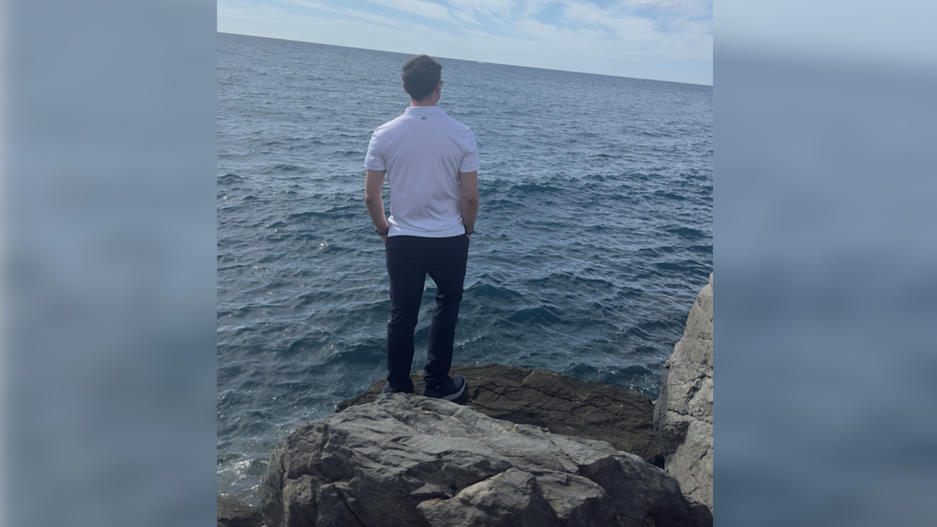 Ein Mann steht mit dem Rücken zur Kamera auf einem Felsen und schaut aufs Meer. 