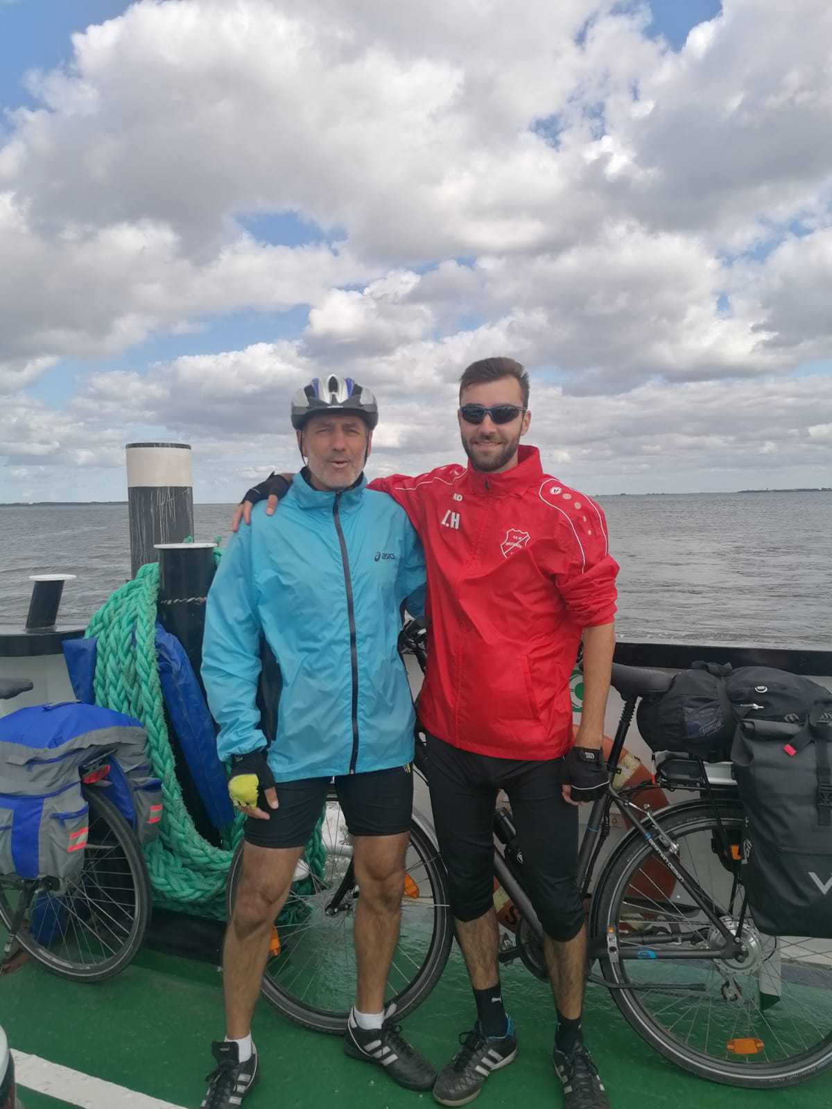 Zwei Männer stehen mit Fahrradbekleidung auf einer Fähre am Wasser. Der ältere Mann links trägt eine blaue Jacke und einen Fahrradhelm, der jüngere Mann rechts eine rote Sportjacke und Sonnenbrille. Beide haben ihre Fahrräder dabei, die mit Gepäcktaschen beladen sind. Im Hintergrund ist eine weite Wasserfläche unter einem teils bewölkten Himmel zu sehen.