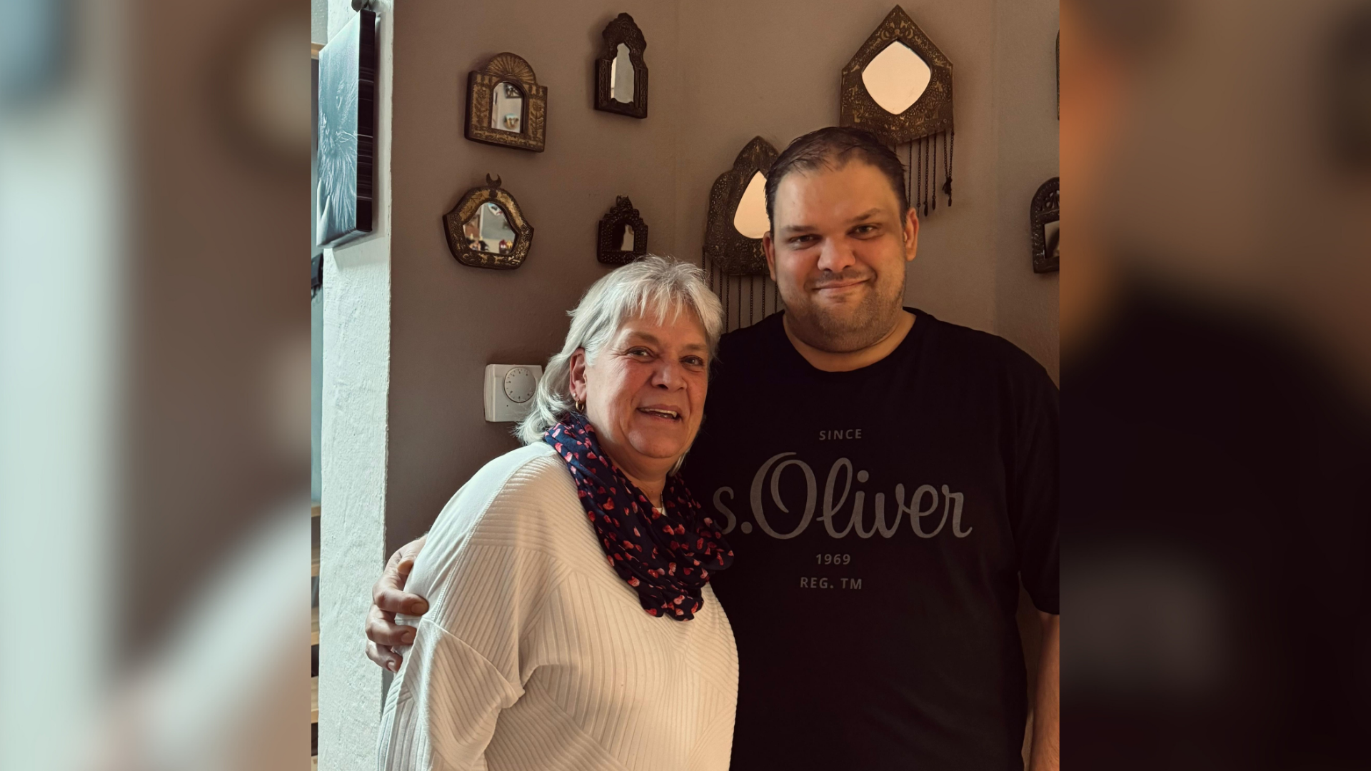 Ein Mann und eine Frau stehen Arm in Arm vor einer braun gestrichenen Wand und lächeln in die Kamera. 