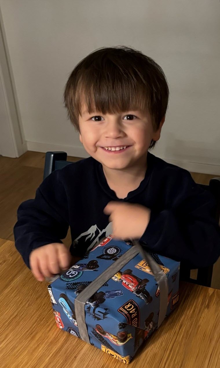 Ein kleiner junge mit dunkeln Haaren sitzt an einem Tisch und packt ein Geschenk aus.