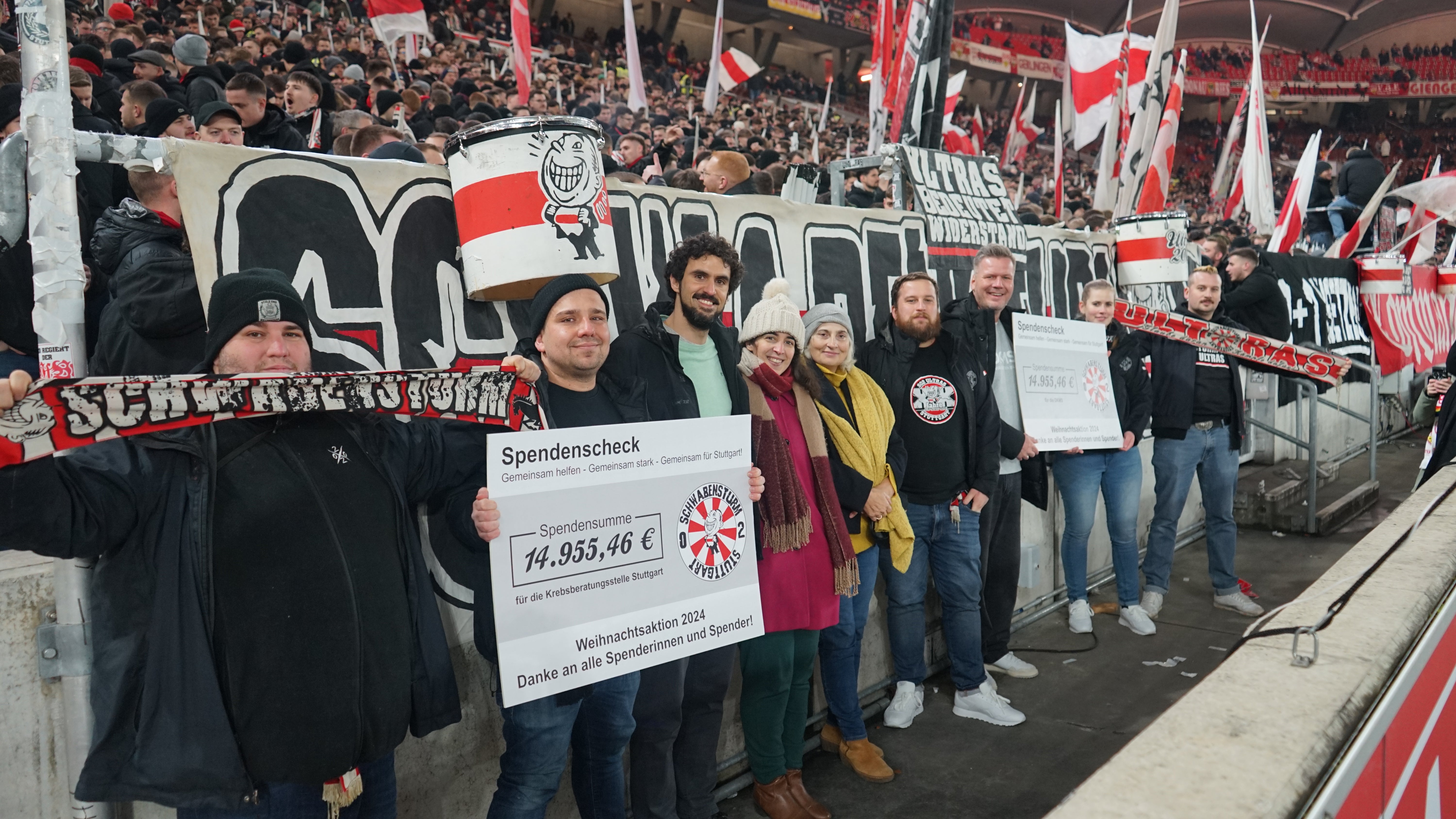 Mehrere Menschen stehen aufgereiht vor der Fantribüne im Stuttgarter Stadion, halten Schals hoch und zwei symbolische Spendenschecks.