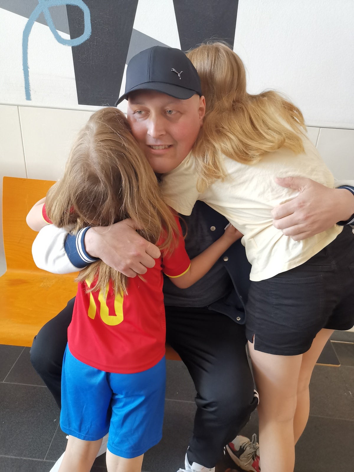 Ein Mann mit schwarzer Baseballkappe sitzt auf einer orangen Bank und umarmt zwei Kinder. Eines der Kinder trägt ein rotes Trikot mit der Nummer 10, das andere ein helles T-Shirt. Beide Kinder sind von hinten zu sehen. Im Hintergrund ist eine helle Wand mit abstrakten schwarzen und blauen Formen.