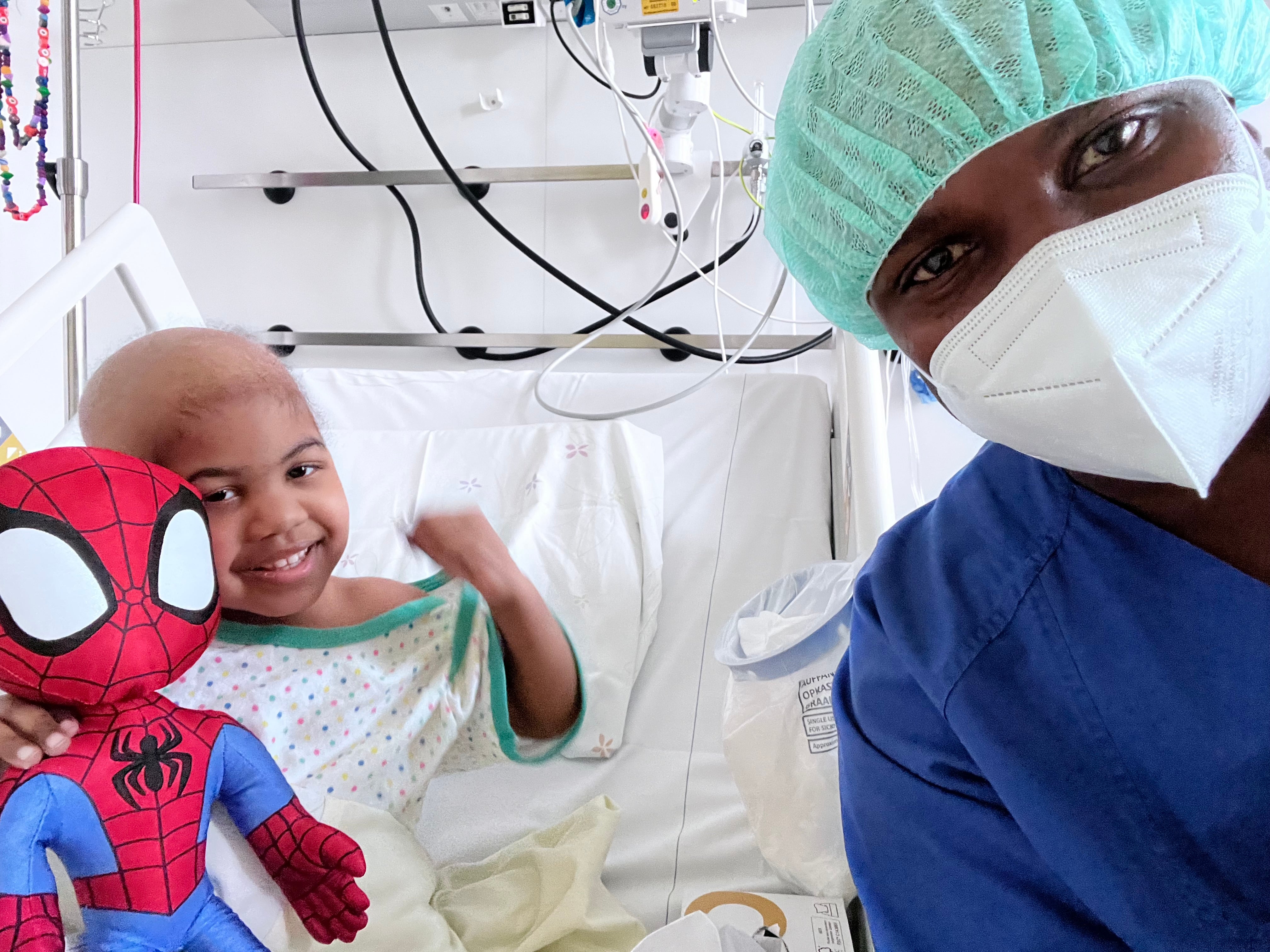 ein Kind liegt mit einer Spiderman Puppe in einem Krankenhausbett, im Vordergrund sieht man einen Mann mit OP Kleidung und Maske