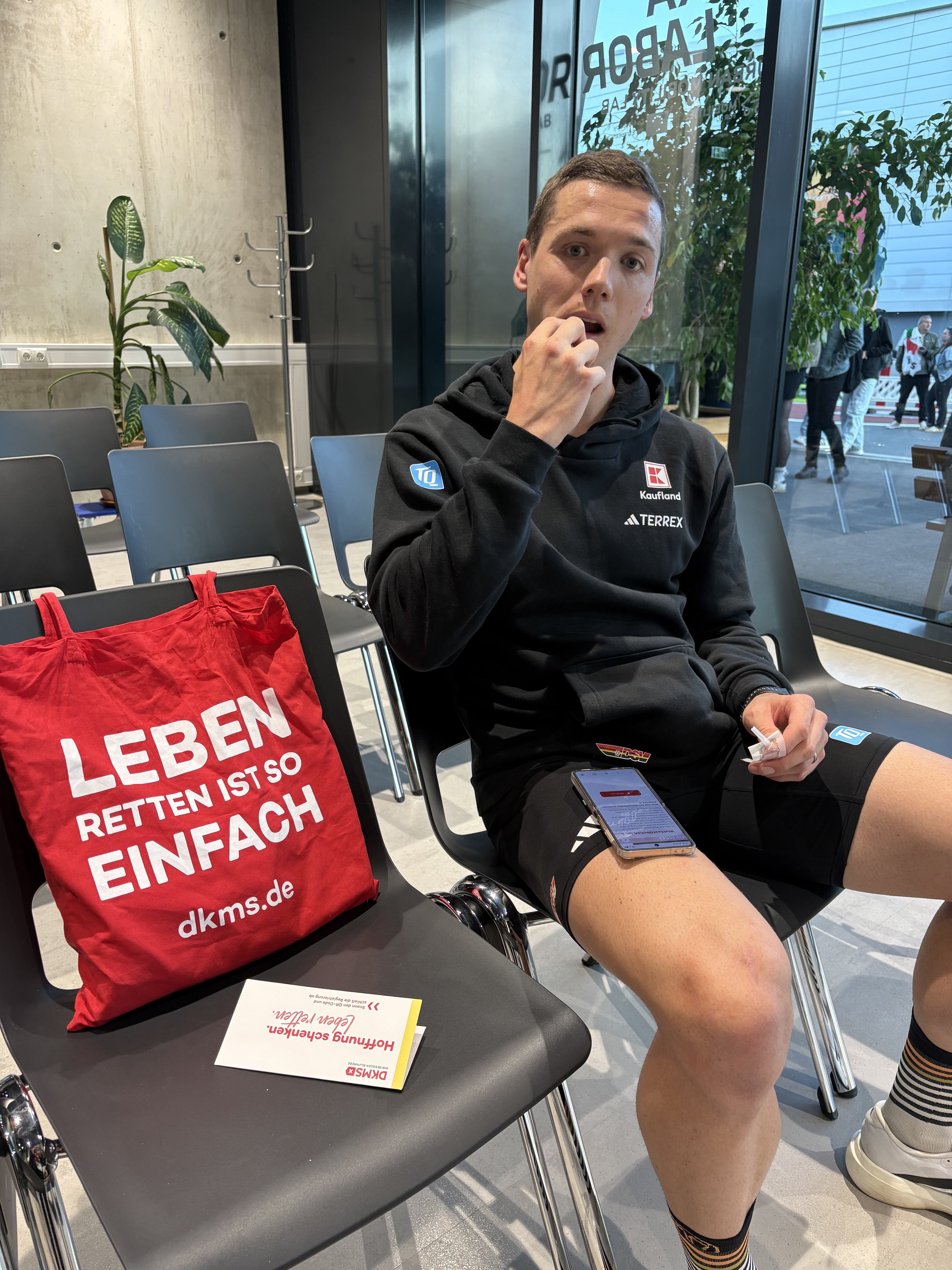 Ein Mann, der auf einer schwarzen Stuhlreihe sitzt. Neben ihm liegt ein roter DKMS Beutel.