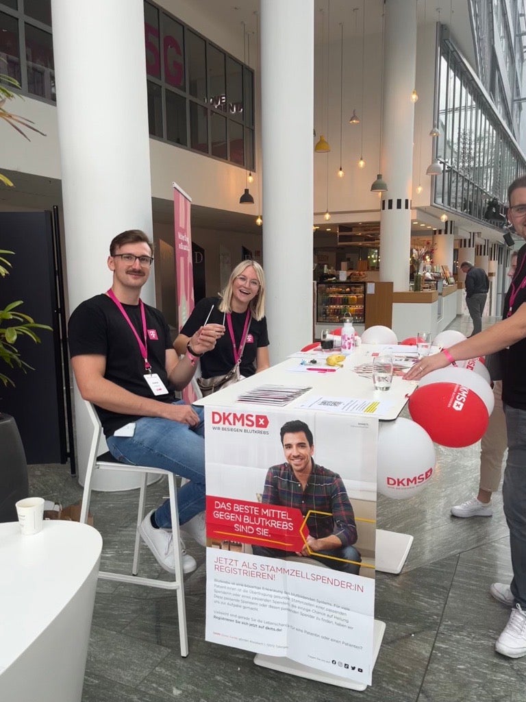 Zwei Personen mit pinken DKMS-Lanyards sitzen an einem DKMS-Informationsstand in einem modernen, hellen Gebäude mit hoher Glasfassade. Beide halten Wattestäbchen in der Hand und lächeln in die Kamera. Auf dem Tisch liegen Registrierungsunterlagen und DKMS-Materialien, darunter rote Luftballons. Im Vordergrund hängt ein Plakat mit dem Slogan „Das beste Mittel gegen Blutkrebs bist du“ und dem Aufruf zur Stammzellspende. Eine dritte Person steht am rechten Bildrand, teilweise im Anschnitt.
