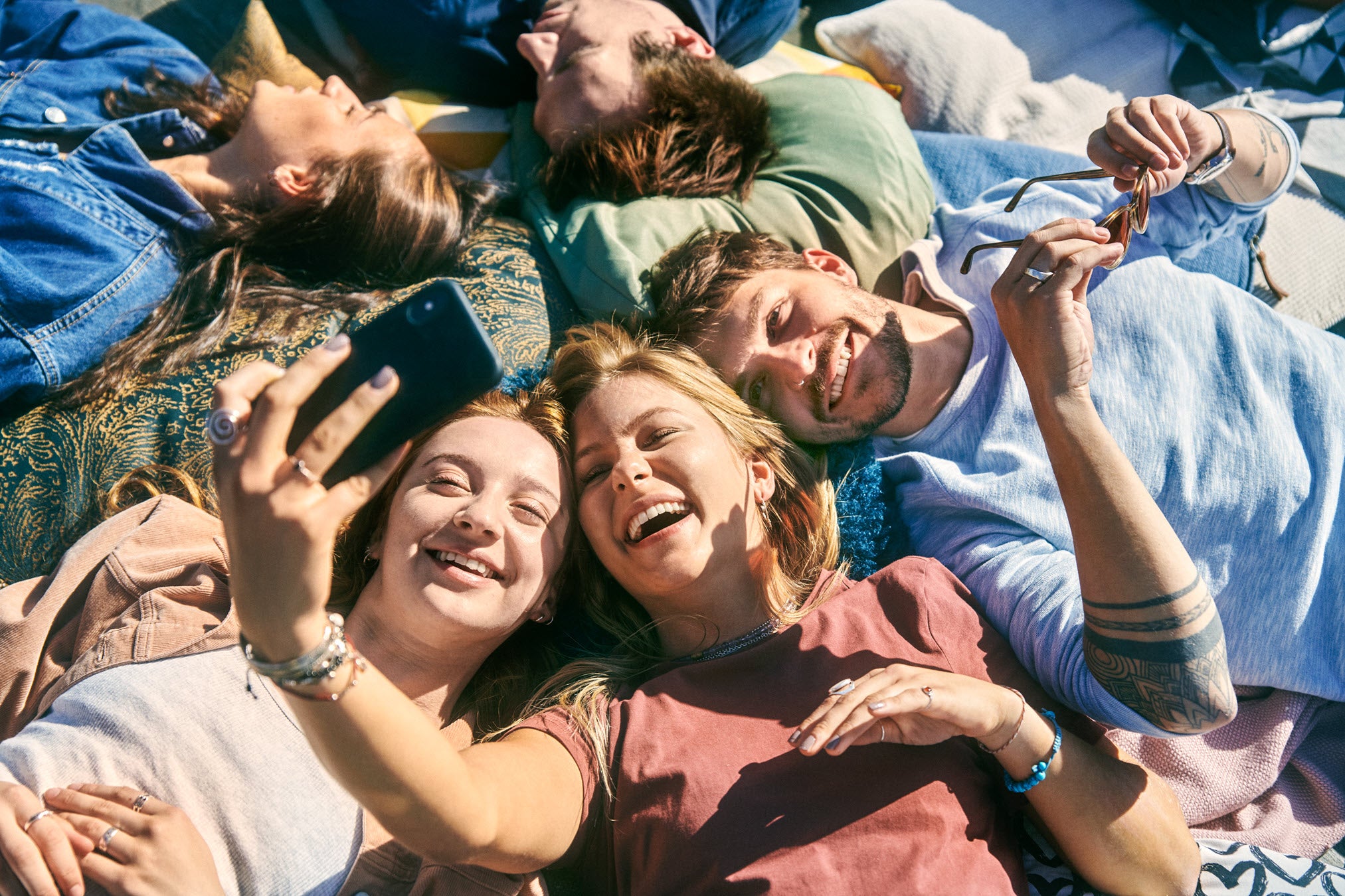 Mehrere junge Erwachsene liegen entspannt auf Decken im Freien und machen gemeinsam ein Selfie. Im Vordergrund halten zwei Frauen ein Smartphone und lachen in die Kamera. Weitere Personen liegen um sie herum, einige mit geschlossenen Augen oder lächelnd. Die Stimmung ist fröhlich und locker.
