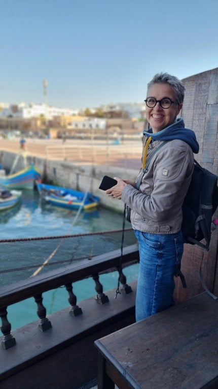 Eine Frau steht auf einem Holzschiff mit einem Gewässer und Booten im Hintergrund.