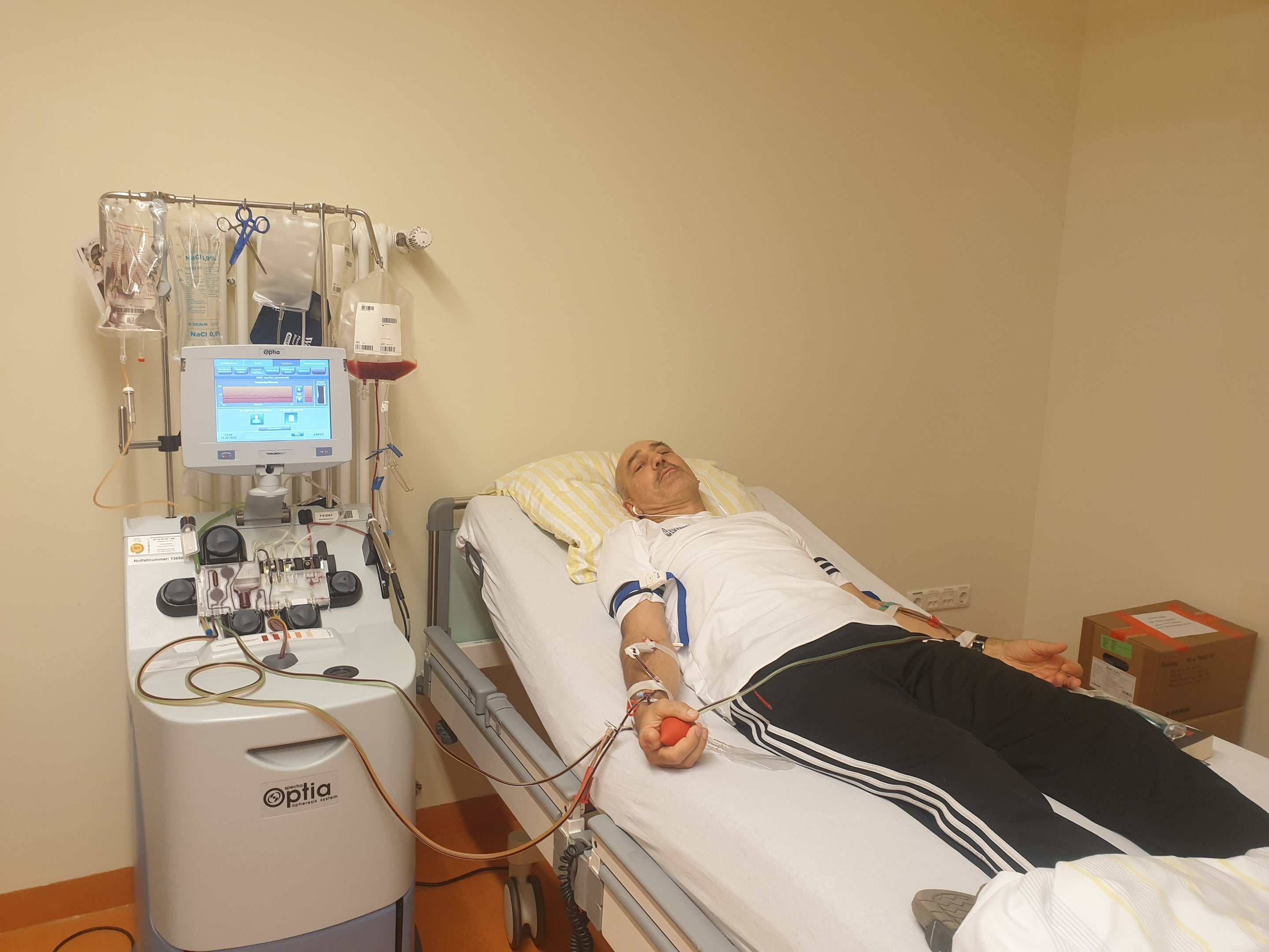Ein Mann liegt während einer peripheren Stammzellentnahme entspannt in einem Klinikbett. Er hält einen roten Gummiball in der Hand, während sein Blut über einen Schlauch in eine Apheresemaschine geleitet wird. Neben dem Bett steht das medizinische Gerät mit einem Bildschirm, mehrere Infusionsbeutel hängen daran.