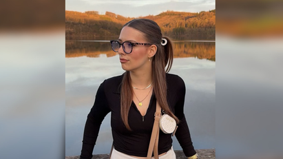 Eine Frau trägt Brille und steht vor einem See. 
