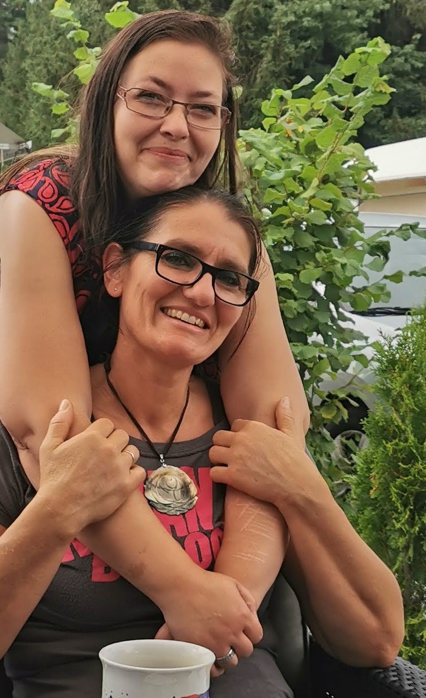 Zwei Frauen sitzen eng beieinander im Freien. Die Frau im Hintergrund umarmt die Frau im Vordergrund von hinten über die Schultern. Beide tragen Brillen und lächeln. Im Vordergrund steht ein weißer Becher auf einem Tisch. Im Hintergrund sind grüne Pflanzen und Bäume zu sehen.