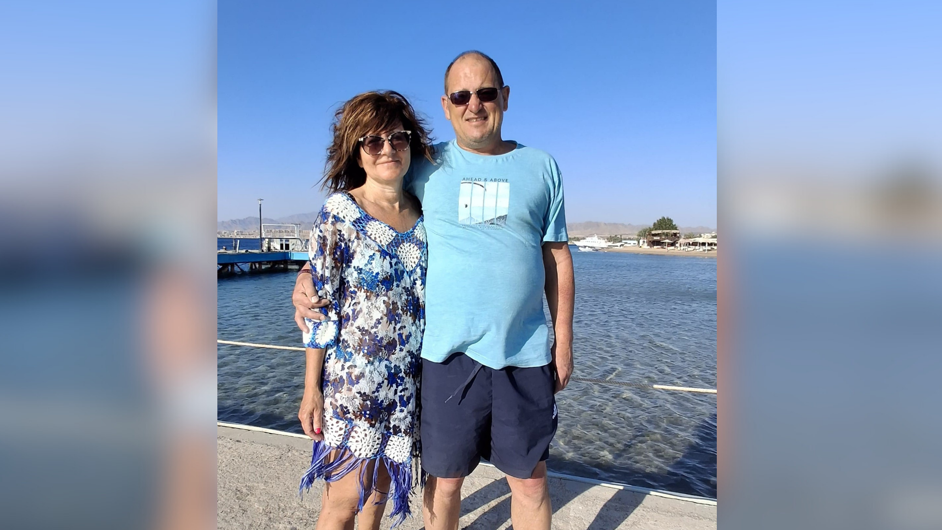 Eine Frau und ein Mann stehen Arm in Arm und sommerlich bekleidet an einem Pier.