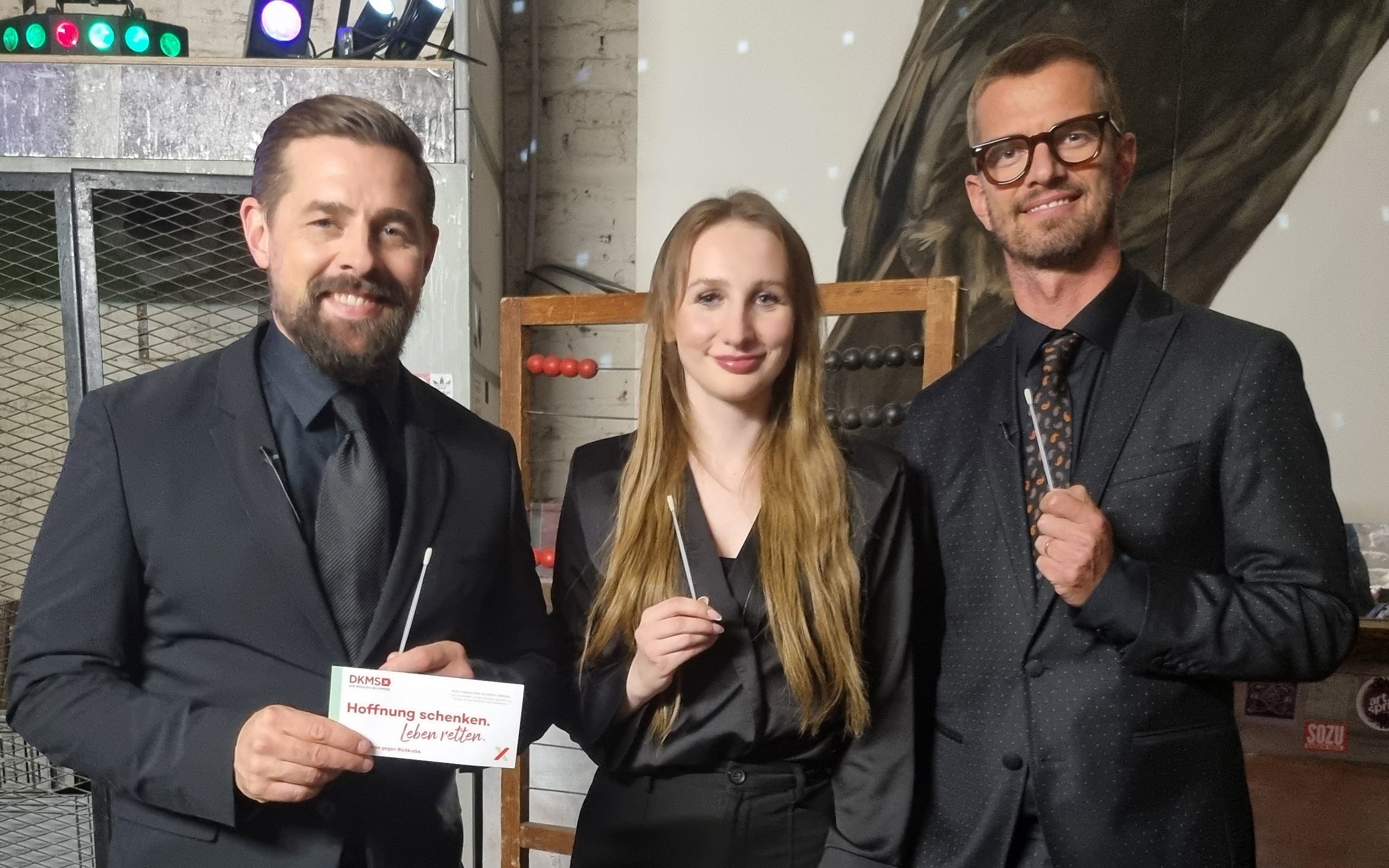 Zwei Männer und eine Frau stehen nebeneinander und blicken lächelnd in die Kamera. Alle drei tragen dunkle Kleidung. Die Frau und der Mann rechts halten jeweils ein Wattestäbchen in der Hand. Der Mann links hält zusätzlich ein DKMS-Kärtchen mit der Aufschrift „Mund auf. Stäbchen rein. Spender sein.“. Im Hintergrund sind bunte Lichter und metallische Regale zu sehen.