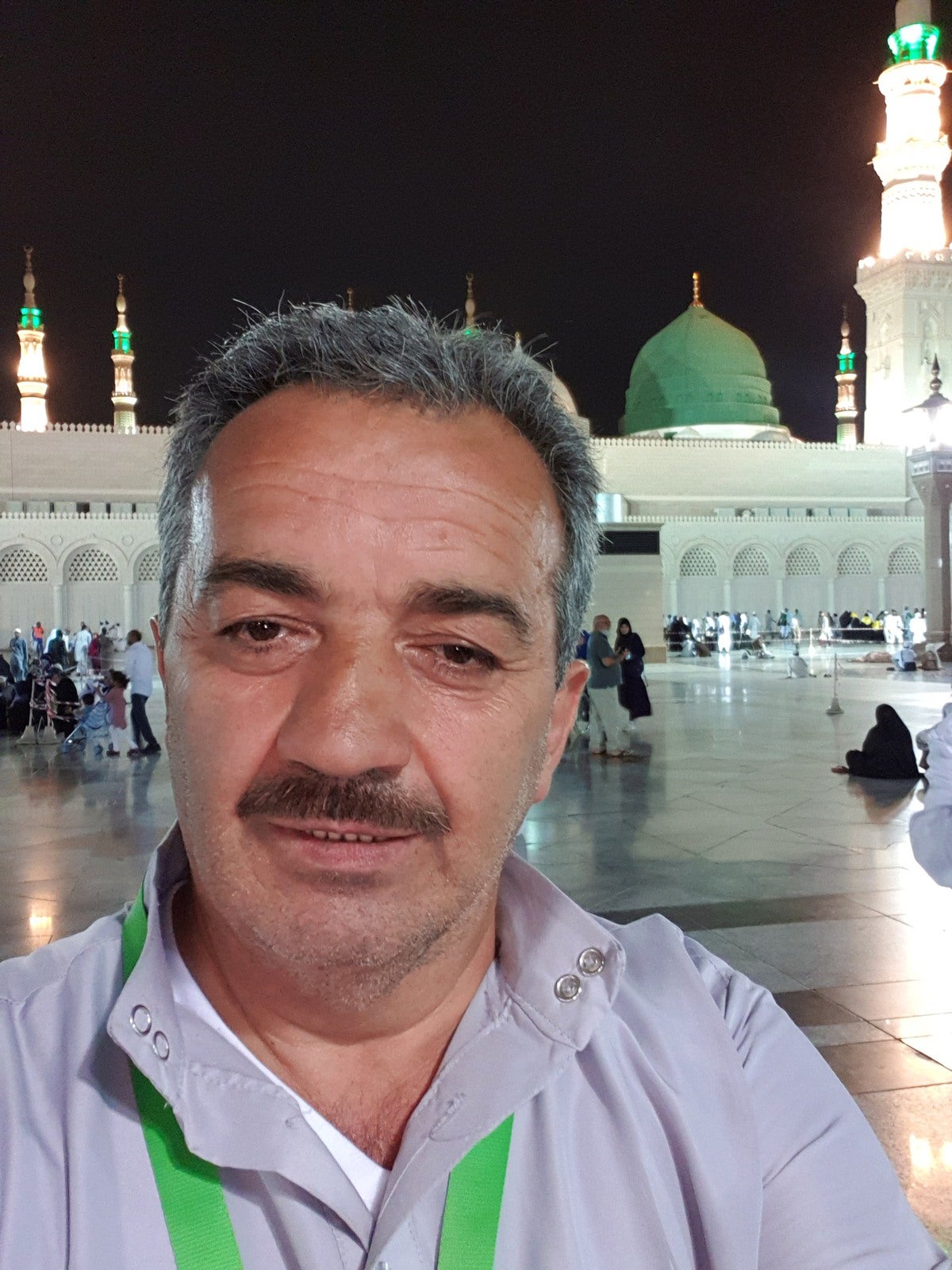 Ein Mann macht ein Selfie auf einem hell erleuchteten Platz vor der Prophetenmoschee in Medina. Im Hintergrund sind die grüne Kuppel und mehrere beleuchtete Minarette zu sehen. Der Mann trägt ein helles Hemd mit einem grünen Umhängeband und blickt direkt in die Kamera.