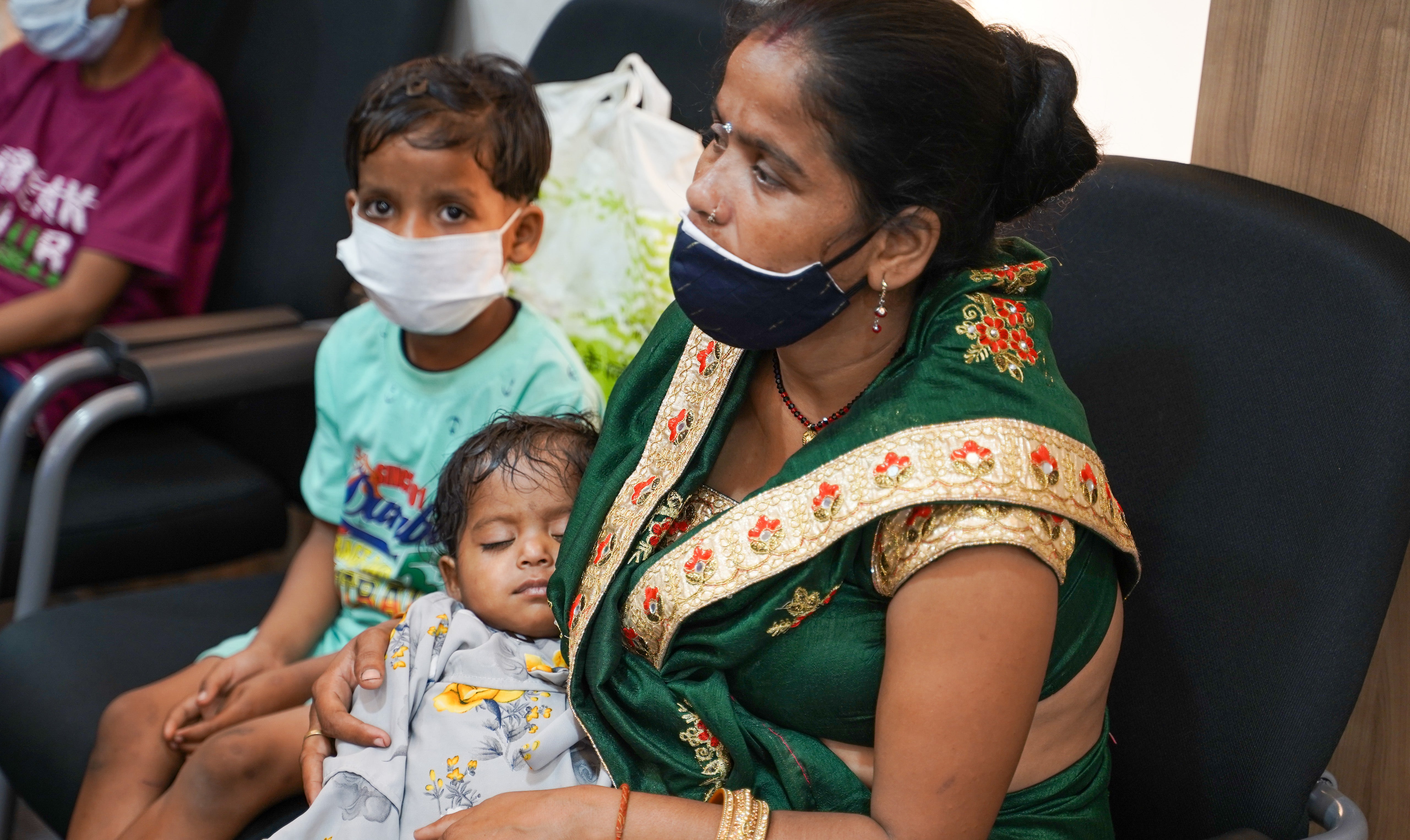 Eine Frau hält ein Baby, neben ihr sitzt ein kleines Kind. Sie tragen Masken