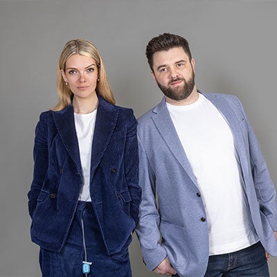 A photo of Celia Chartres-Aris and Jamie Shields. Celia stands on the left, she has long blonde hair and wears a blue suit. Jamie stands on the right, he has short brown hair and a beard. He wears a light blue blazer.