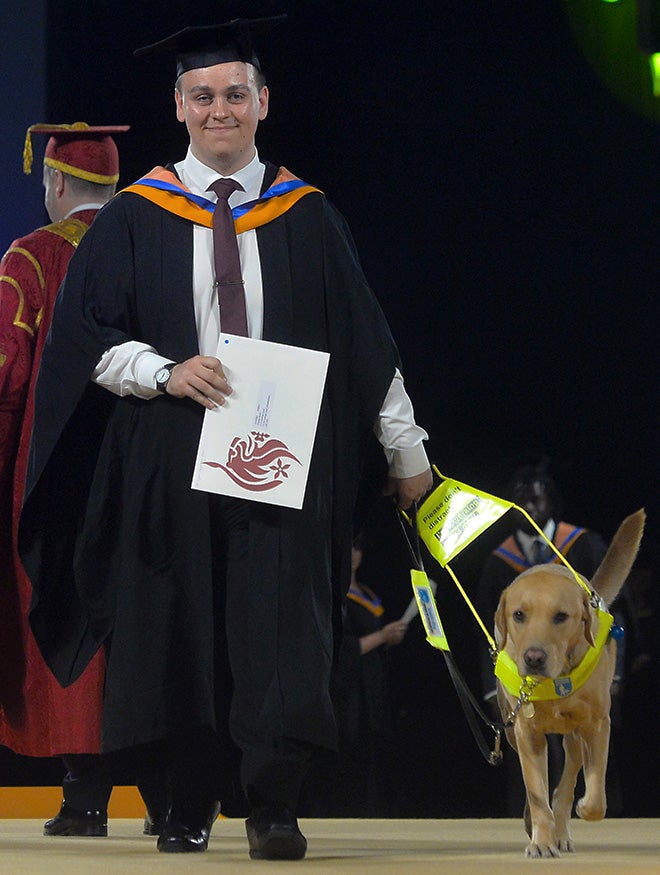 A smiling young man wearing graduation robes holding a certificate and walking with his guide dog in a hall