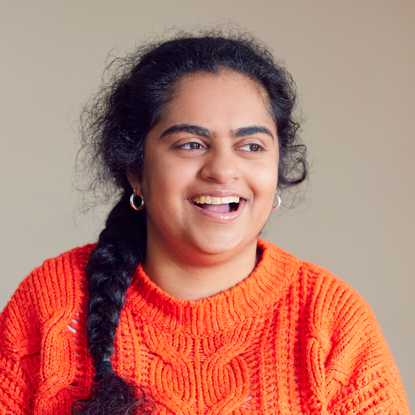 Portrait photo of a woman laughing against a plain background. She is wearing a red orange woollen jumper.
