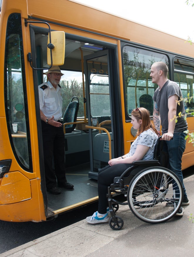 A man pushes a woman in a wheelchair toward the entrance toward a bus where the driver stands in the doorway