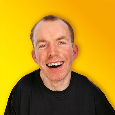A photo of Lee Ridley smiling in front of a yellow background. He wears a black t-shirt. 