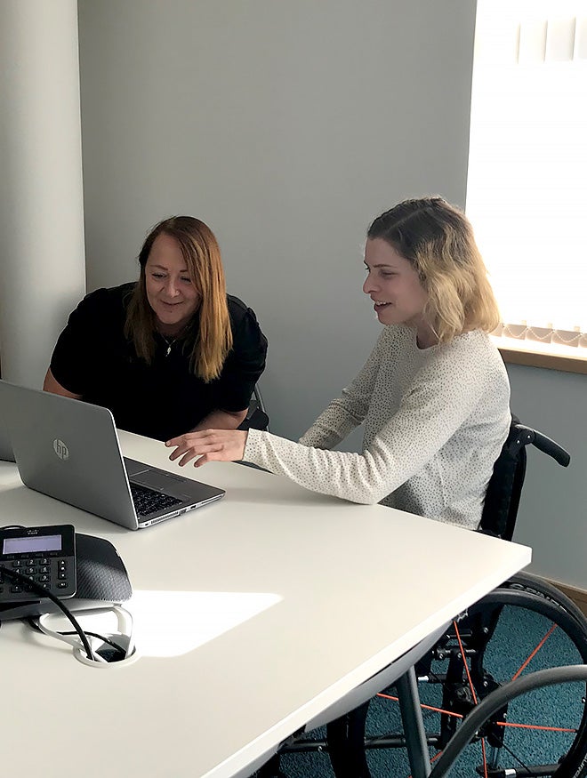 A smiling young woman in a wheelchair with a smiling woman, sitting at a table in an office meeting room looking at a laptop