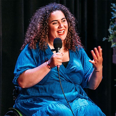 A photo of Chloe van Dalen smiling and holding a microphone. She has dark curly hair and wears a blue dress. 