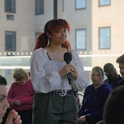 A member of The Assembly stands holding a microphone. They have curly red hair and are wearing a white blouse.
