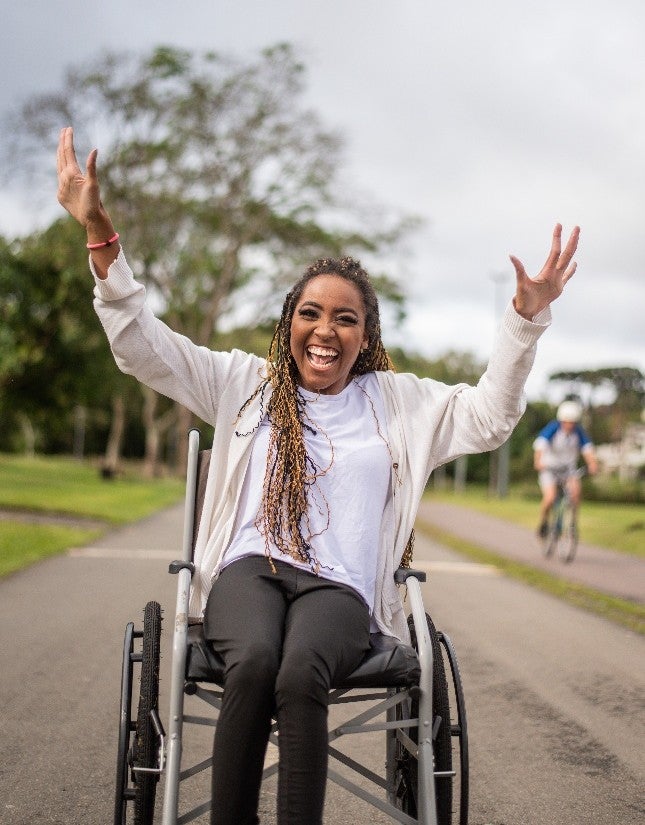 A smiling Black woman in a wheelchair with both arms raised in park path outdoors