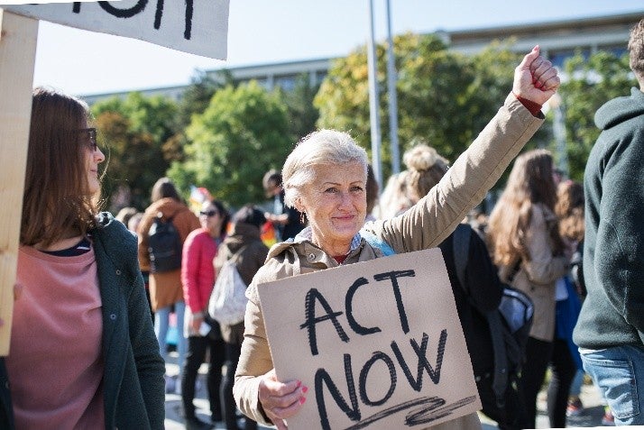 A smiling woman holding a sign that says ACT NOW at a public demonstration