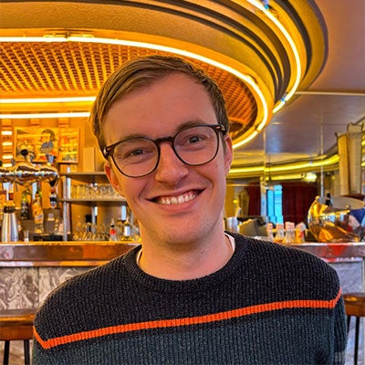 A photo of Drew Hyndman smiling at the camera. He has short hair and wears black-rimmed glasses. He is wearing a black jumper with a red stripe across the chest.