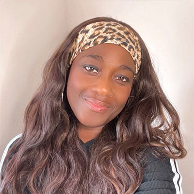 Natalie Diana Busari smiles at the camera. She has long brown hair and is wearing a leopard print headband and a back top with white stripes down the sleeves. 