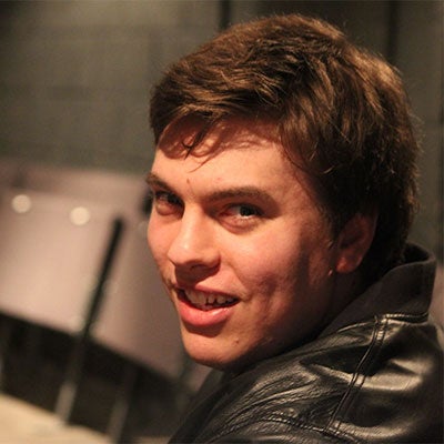 A photo of Alex Taylor smiling. He has short, brown hair and is wearing a black leather jacket. 