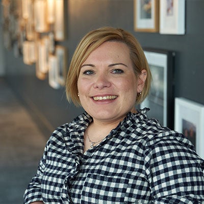 A photo of Sam Tatlow smiling. She has short blonde hair and wears a black and white checked shirt. 
