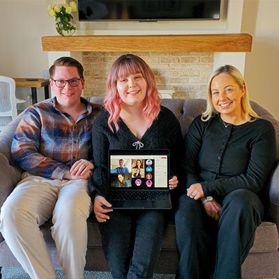 A photo of the Inkfire team. Three team members sit on a sofa, showing a laptop with other team members on a video call.