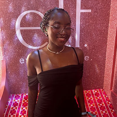 Charis Townsend smiles in front of a glittery pink background. She wears glasses and a nose ring. She is wearing a black strappy dress. 