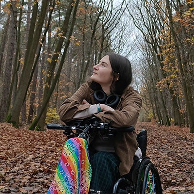 A photo of Zara Beth in the woods. There is a rainbow bandanna tied onto her wheelchair. 