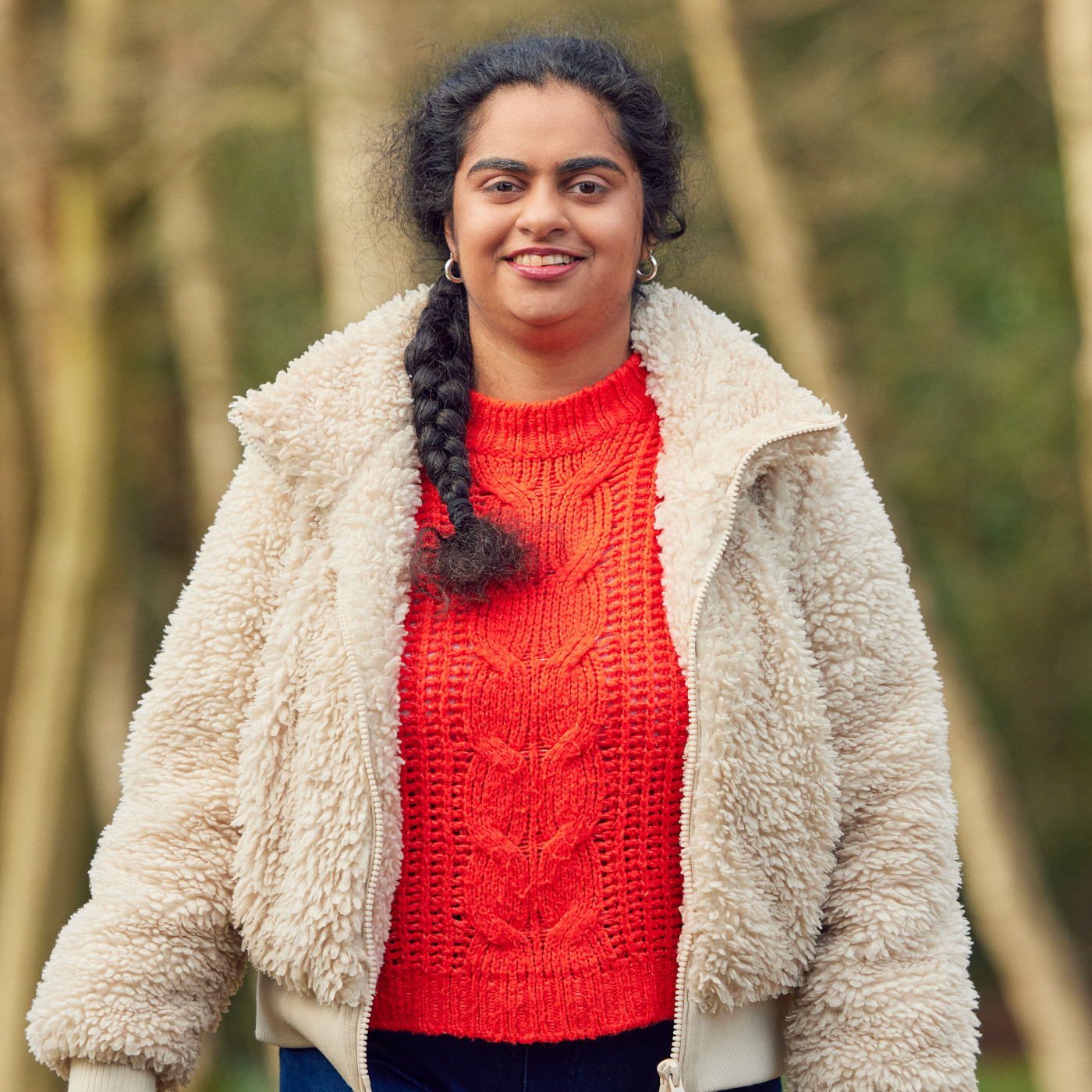 Portrait photo of a smiling woman wearing fur coat and red orange woollen jumper standing outside in woodland