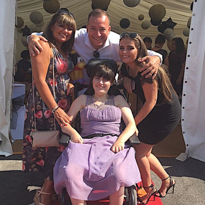 A smiling young woman in a wheelchair with her mum, dad and big sister outside on a sunny day