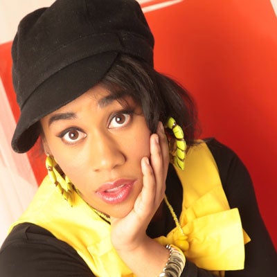 A photo of Variety D wearing a black cap, yellow earrings, and a yellow top. 